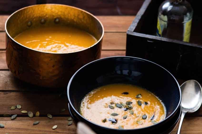 Hier erkennt man eine Schüssel und ein Topf mit einer köstlichen, orangenen Kürbissuppe, diese ist mit Kürbiskernen garniert. Ein Löffel liegt zum essen bereit.