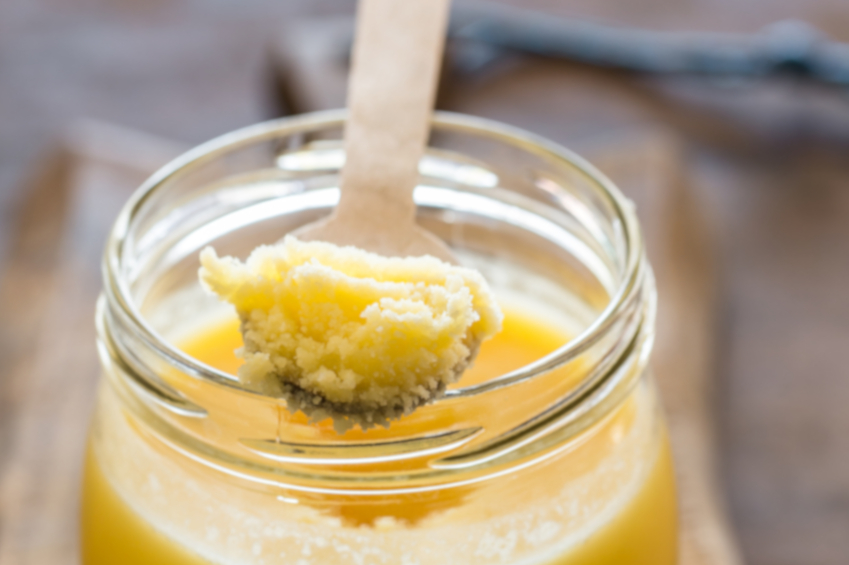 Man erkennt ein Glas mit Ghee. Auf dem Rand liegt ein Löffel, der voll ist mit Ghee.