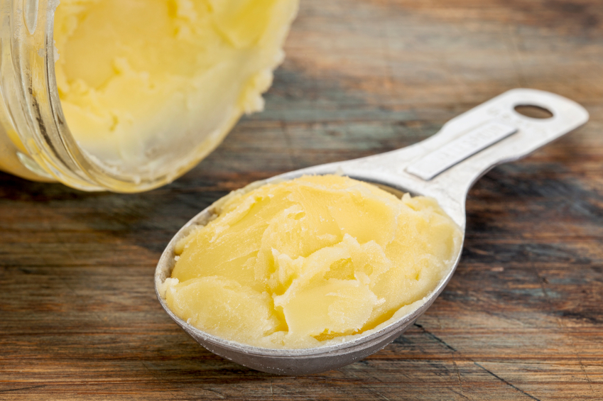 Auf einem strukturierten Holztisch liegt ein offenes Glas mit Ghee. Davor liegt ein Messlöffel auf den Ghee gestrichen ist.