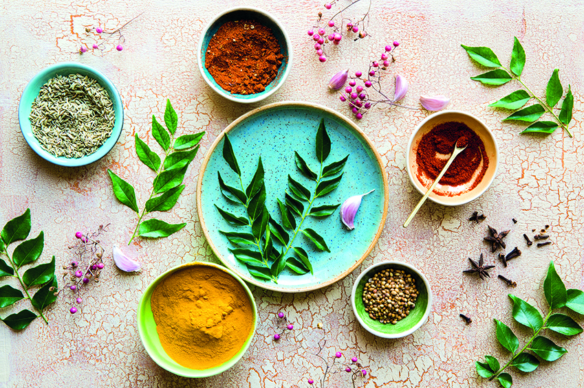 Verschiedene Kräuter, Gewürzmischungen und Gewürze in Schälchen. Dazu liegen frische Curry Blätter, Nelken und Knoblauch